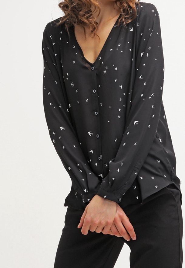 Blouse noire à manches longues, col en V et fermeture par boutons, ornée d'un imprimé d'oiseaux blancs sur l'ensemble, en tissu lisse et léger.