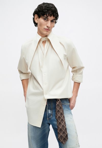 Off-white button-up shirt with an oversized collar, featuring a gathered front design and rolled sleeves, paired with distressed denim and a patterned tie.