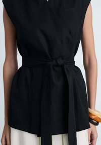 Top avvolgente in linom nero con maniche corte e vita annodata, caratterizzato da una vestibilità rilassata e una superficie liscia. Dettaglio della piccola tasca frontale visibile.