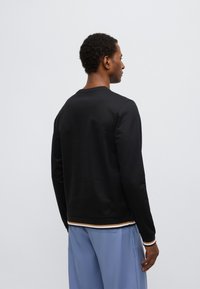 Juodas megztinis ilgomis rankovėmis ir apvalia iškirpte, turintis tekstūruotą paviršių ir austomą kraštą su kontrastingomis baltomis bei rudiomis juostelėmis.