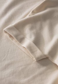 Tessuto di cotone beige con il bordo della manica piegato, che mostra una texture liscia e cuciture sottili. Materiale morbido e leggero disposto in piano.