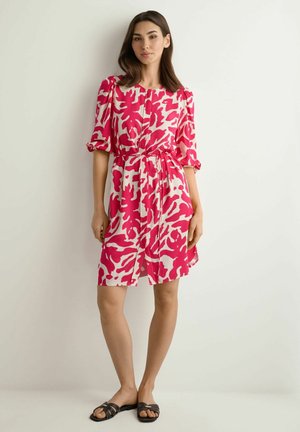 Kobieta stoi przy białej ścianie, ubrana w różowo-białą sukienkę w wzory, o długości do kolan, z półdługimi rękawami i czarne płaskie sandały.