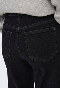 Gros plan sur le dos d'une personne portant un jean en denim noir avec deux poches arrière et un haut gris rentré.