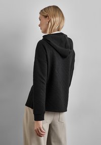 Felpa nera con un motivo geometrico a texture, dotata di cappuccio fisso e un fit rilassato. Indossata con pantaloni casual chiari.