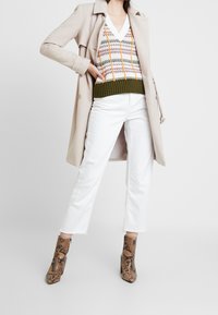 Trench beige sobre un suéter estampado con ribetes verdes, jeans blancos y botas de tobillo de estampado de serpiente marrón con tacón alto.