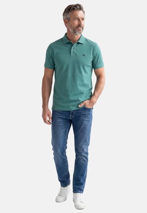 Mężczyzna z siwymi włosami i brodą, ubrany w zieloną koszulkę polo, niebieskie dżinsy i białe trampki, stojący z jedną ręką w kieszeni.