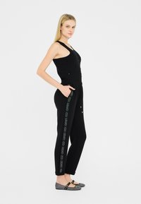 Zwarte sportieve jumpsuit met een aansluitend ontwerp en versierde voorkant, gecombineerd met zwarte broek met een logo langs de zijkant en platte schoenen.