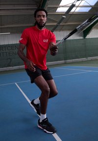 Polo rosso con logo, pantaloni corti neri e scarpe da ginnastica nere con accenti bianchi. Giocatore in azione su un campo da tennis indoor blu.