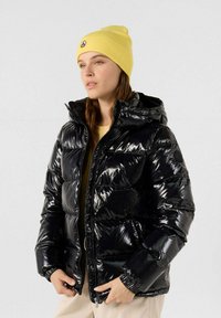 Giacca a vento nera con finitura lucida, chiusura con zip e cappuccio. Indossata sopra una maglietta gialla. Berretto giallo a maglia con logo in alto.