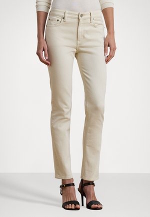 Woman wearing slim beige jeans and black open-toe high heel sandals, standing against plain white background.