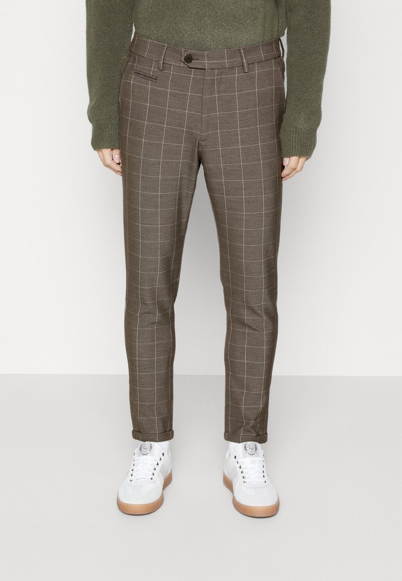 Personne portant un pantalon slim marron à carreaux, retroussé aux chevilles, assorti à des baskets blanches et un pull vert à manches longues.