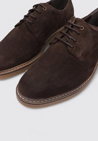 Zapatos de ante marrón con puntera redondeada, diseño de cordones y costuras claras en contraste. Suela de goma con una textura suave.