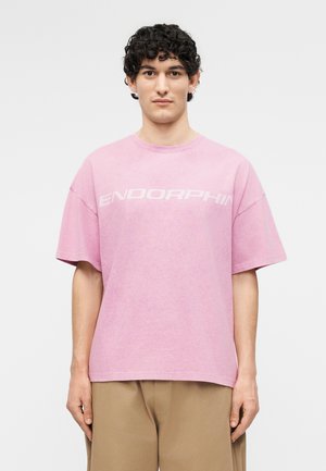 Νεαρός με σγουρά μαλλιά που φοράει μια φαρδιά ροζ μπλούζα με το κείμενο "ENDORPHIN" και μπεζ παντελόνι, στέκεται μπροστά σε απλό ανοιχτό φόντο.