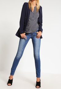 Marineblazer, gemusterte Bluse und enganliegende blaue Jeans; schwarze offene High-Heels komplettieren das Outfit mit einem eleganten, modernen Look.