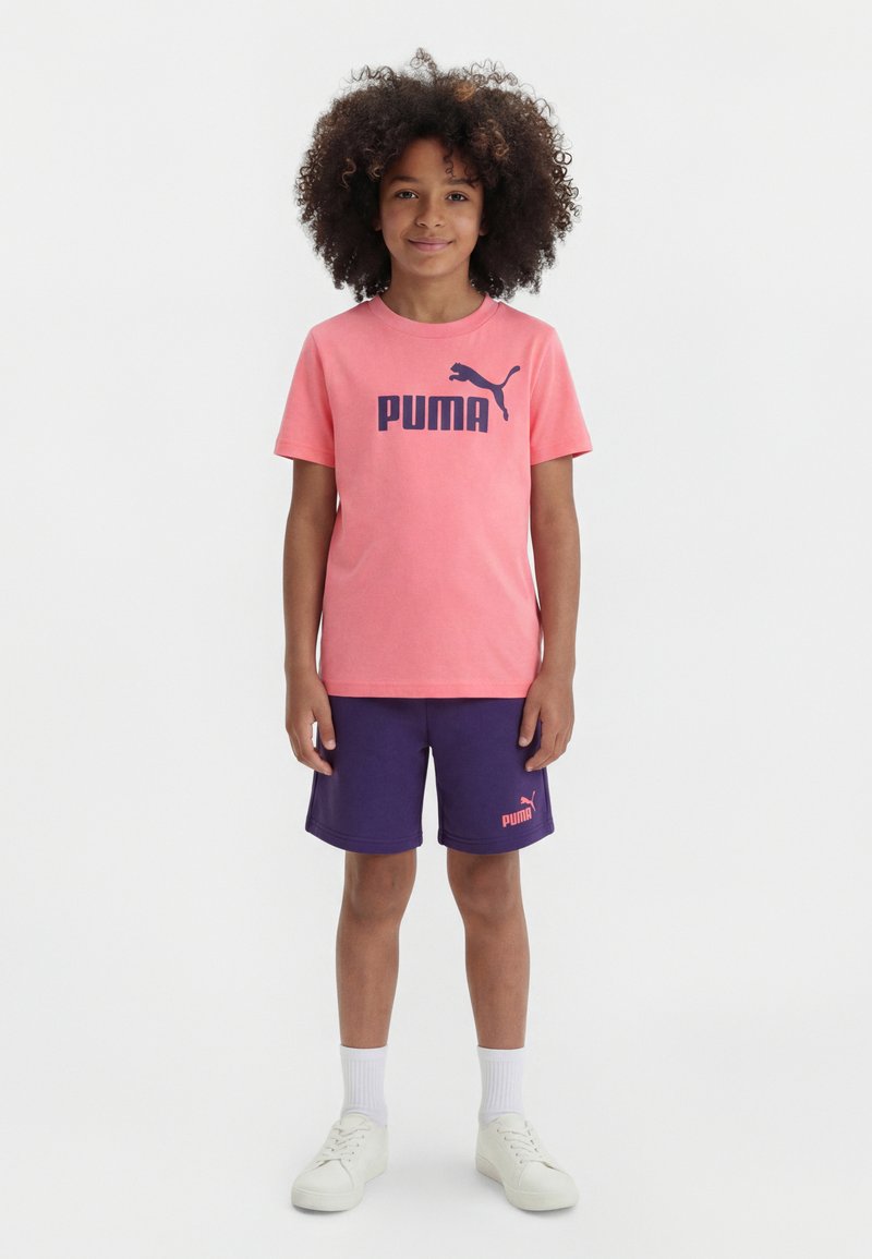 Παιδί με σγουρά μαλλιά όρθιο, που φοράει ροζ μπλουζάκι Puma, μοβ σορτς Puma, λευκές κάλτσες και λευκά αθλητικά παπούτσια σε απλό φόντο.