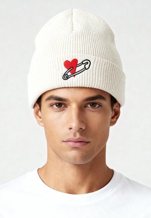 Ribbed white beanie featuring a red heart and safety pin embroidered design. Lightweight knit material with a snug fit.