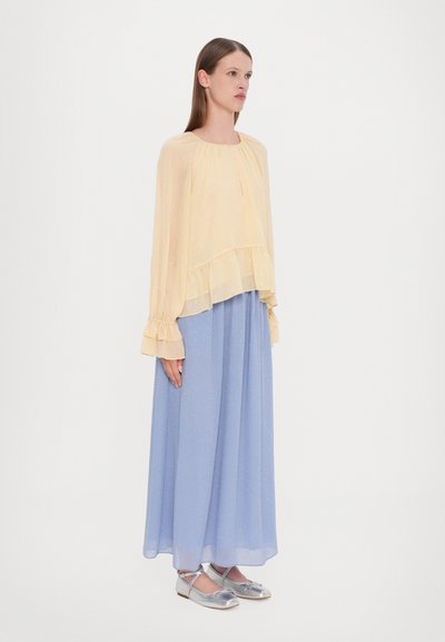 Gele doorzichtige blouse met ruches over een lichtblauwe vloeiende rok. De outfit heeft subtiele sprankeling en zilveren flats.