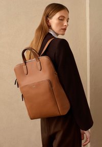 Zaino in pelle marrone chiaro con design minimalista, doppie maniglie e tasca frontale con zip. Superficie texturizzata; dettagli in metallo sulla zip.