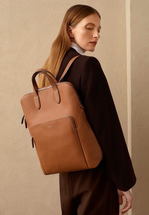 Zaino in pelle marrone chiaro con design minimalista, doppie maniglie e tasca frontale con zip. Superficie texturizzata; dettagli in metallo sulla zip.