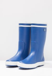 Un par de botas de lluvia de goma azules con ribetes y suelas blancas, de pie sobre una superficie blanca.