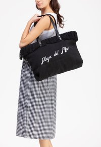 Sort tote bag med frynser i toppen, med hvid tekst "Playa del Mar" og sorte læderhåndtag, båret med en ternet midi-længde nederdel.