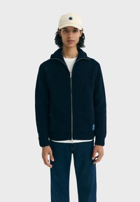 Marineblauwe geribbelde gebreide zip-up jas met hoge kraag, witte onderhemd, en een wolachtige structuur. Gecombineerd met donkerblauwe jeans en een crèmekleurige pet.