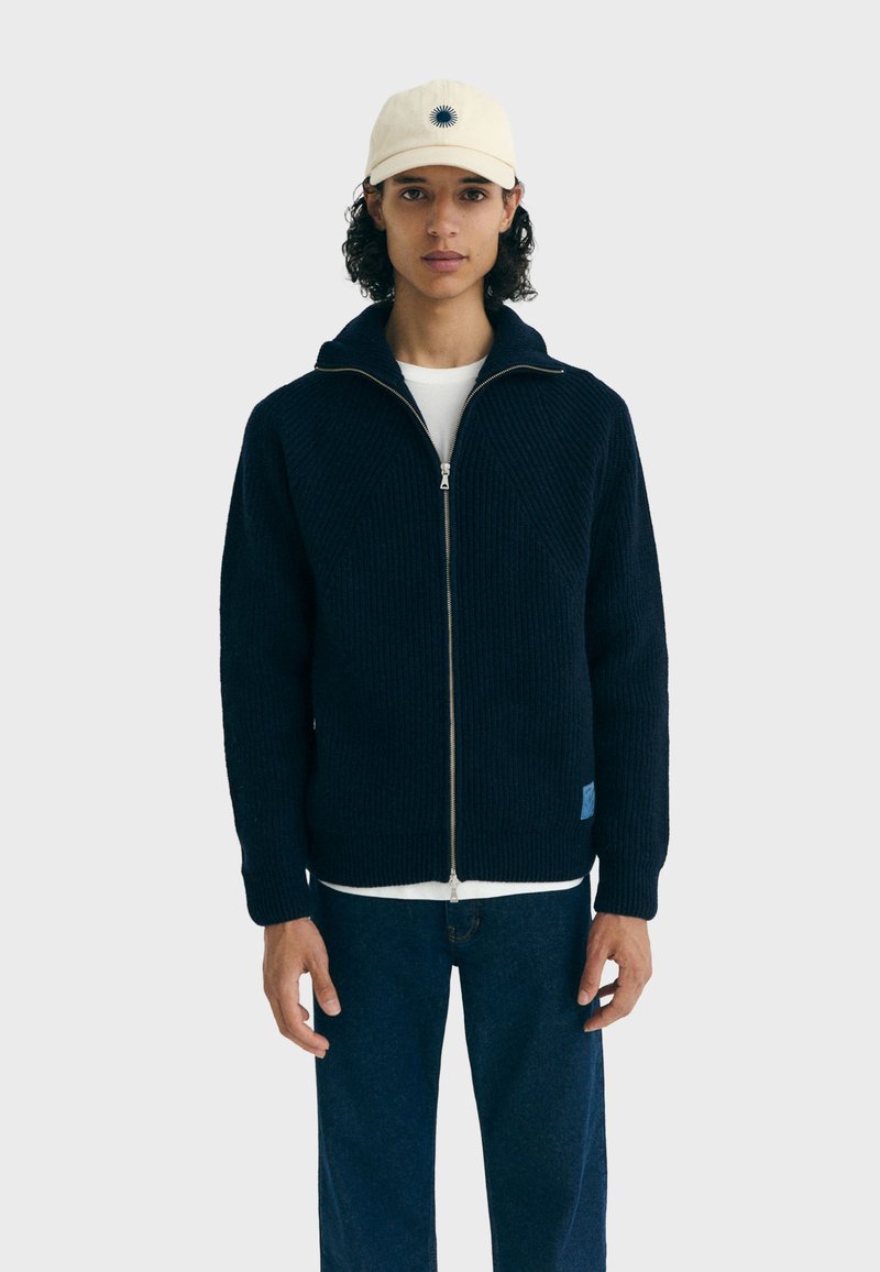Marineblauwe geribbelde gebreide zip-up jas met hoge kraag, witte onderhemd, en een wolachtige structuur. Gecombineerd met donkerblauwe jeans en een crèmekleurige pet.