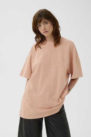 Γυναίκα με καστανά μαλλιά μέχρι τον ώμο φοράει φαρδύ ροδακινί μπλουζάκι και μαύρο παντελόνι με φαρδύ πόδι, ποζάροντας μπροστά σε απλό γκρι φόντο.