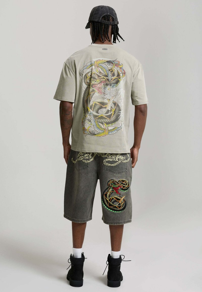 Mann trägt ein beige T-Shirt mit farbenfrohem Schlangen- und Vogeldesign auf dem Rücken, dunkle knielange Shorts mit Schlangenstickerei am rechten Bein.