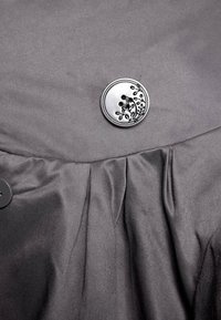 Tessuto grigio dalla texture liscia presenta un bottone rotondo decorativo, metallico con incisioni floreali e due asole. Dettaglio plissettato sotto.