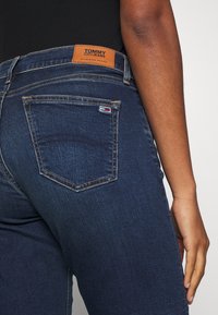 Jeans de mezclilla azul oscuro con una etiqueta de cuero marrón en la cintura, que cuentan con un bolsillo trasero con un pequeño logotipo, mostrando un estilo clásico de cinco bolsillos.
