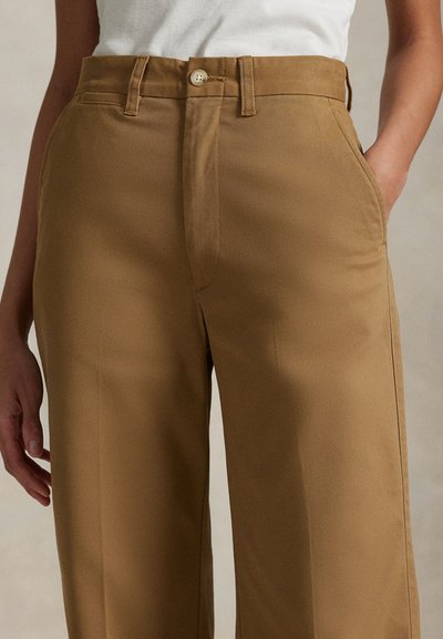 Személy magas derekú, egyenes szárú, teve színű nadrágot visel oldalán zsebekkel, hozzá egy behúzott fehér felsővel.