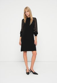 Vestido negro con una silueta ajustada y mangas largas de malla con lentejuelas. Las mangas cuentan con puños fruncidos y la tela tiene una textura suave.