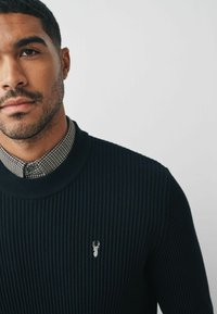 Maglione a coste navy con scollatura rotonda e un piccolo emblema ricamato. Sotto, si intravede una camicia a quadri con colletto.