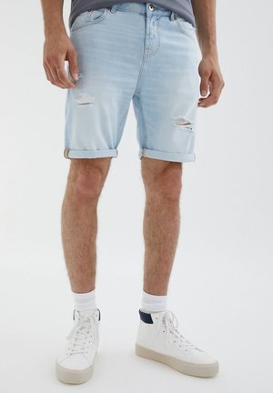 Shorts en denim bleu clair à revers retroussés, présentant des détails usés et une coupe décontractée, associés à des baskets montantes blanches et des chaussettes blanches.