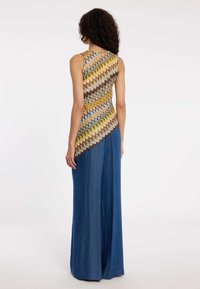 Vrouw met krullend haar draagt een mouwloos topje met zigzagmotief in bruine, gele en blauwe tinten en een brede blauwe broek, staand met de rug naar de camera.