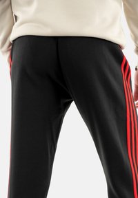 Pantaloni jogger neri con tre strisce verticali rosse lungo i lati, realizzati in un tessuto morbido con una consistenza liscia e una vestibilità rilassata.