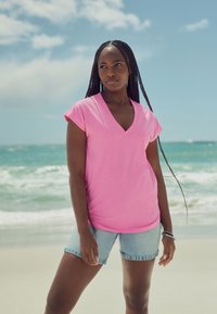 Roza majica s V-izrezom i kratkim rukavima u obliku kapica, uparena s traper kratkim hlačama. Majica ima glatku teksturu i opušteni kroj, prikladna za toplo vrijeme.