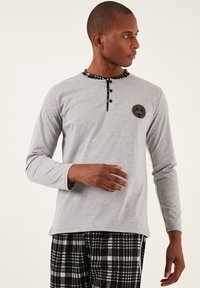 Cămașă gri cu mânecă lungă, cu guler pătratelor negre și placket cu nasturi, având un logo circular pe piept, asortată cu pantaloni de pijama cu carouri.