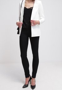Bílý blejzr, černý krajkový top, černé skinny džíny a černé lodičky s špičatou špičkou. Blejzr má klopový límec a štíhlý střih.