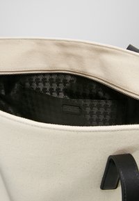 Torba płócienna w kolorze kremowym z czarną podszewką w wzór houndstooth. Posiada górny zamek i czarne skórzane uchwyty. Wnętrze zawiera metkę.