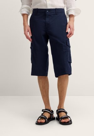 Mann, der marineblaue kniehohe Cargo-Shorts, weiße hochgekrempelte Hemdärmel und schwarze Sandalen trägt, steht vor einem schlichten Hintergrund.