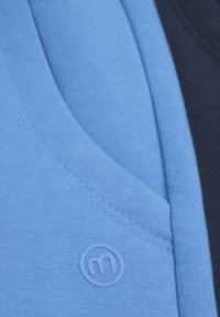 Blauer Stoff mit feiner Textur, der ein kreisförmiges Logo in einem helleren Blauton aufweist. Das Design umfasst einen kontrastierenden dunkelblauen Abschnitt.