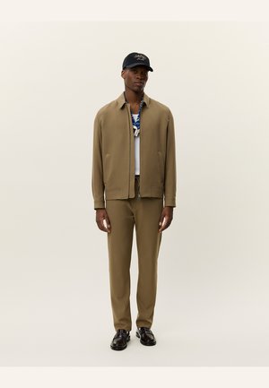 Braune Zip-Jacke über einem weißen Hemd, kombiniert mit braunen Hosen. Dazu eine marineblaue Mütze und schwarze Schuhe. Einfaches, elegantes Design mit einer glatten Textur.