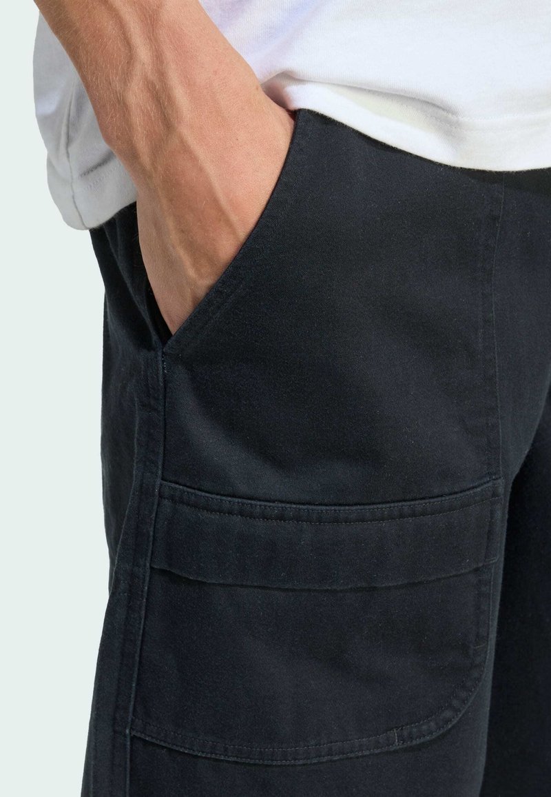 Hand in een zwart cargopantszak met klep, gedragen met een wit shirt.