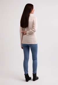 Beige gestrickter Pullover mit geripptem Saum, kombiniert mit hellblauen Skinny Jeans und schwarzen Ankle Boots. Modell von hinten betrachtet. Langes, glattes Haar.