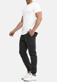 Weißes Kurzarm-T-Shirt, kombiniert mit schwarzen Cargo-Hosen mit seitlichen Taschen und elastischen Bündchen, getragen mit weißen Sportschuhen.