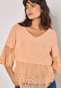 Pfirsichfarbene bestickte Bluse mit V-Ausschnitt, kurzen schwingenden Ärmeln, schichtweisem Design und gewelltem Saum mit floralen Mustern.