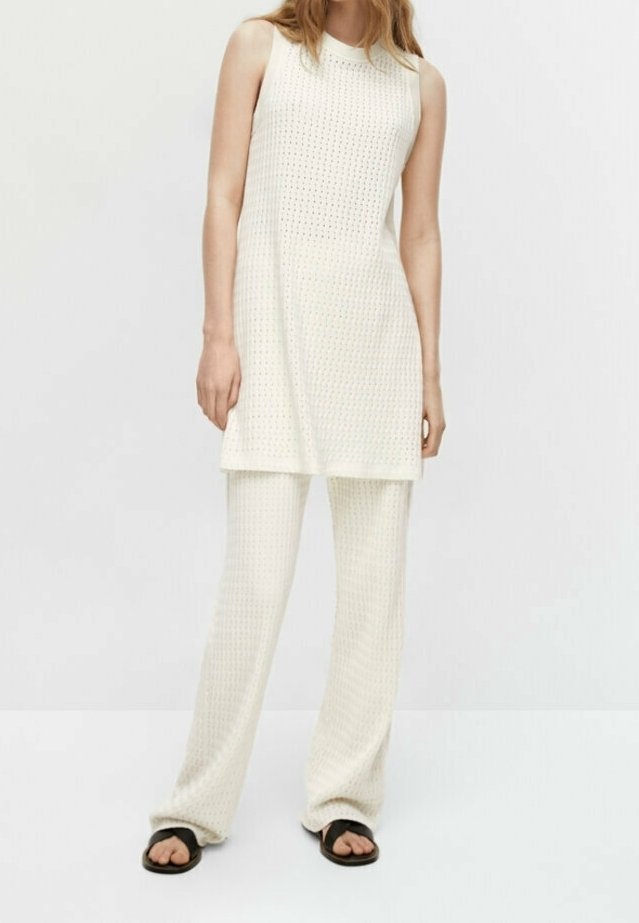 Femme portant une tunique blanche en maille sans manches et un pantalon large assorti avec des sandales noires à bout ouvert, debout devant un fond uni.