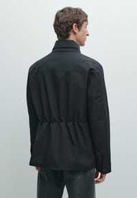 Giacca nera con colletto alto, vita aderente e maniche lunghe. Texture liscia con cuciture minime e polsini abbottonati.